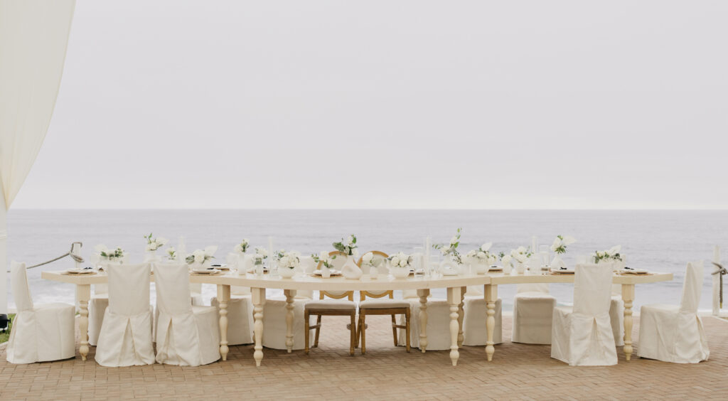 FOTOGRAFIA DEL MONTAJE EN DONDE SE VE LA DECORACION DE LA MESA IMPERIAL, FLORES, COPAS, PLATO Y BAJO PLATO JUNTO CON EL MENU IMPRESO, CUCHARA, TENEDOR Y CUBIERTO. CON EL MAR Y EL FONDO.