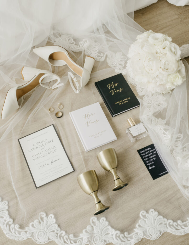 FOTOGRAFIA DE ZAPATOS, ANILLOS, INVITACION, PERFUME Y VOTOS PUESTOS CUIDADOSAMENTE SOBRE EL VELO Y AL LADO EL RAMO