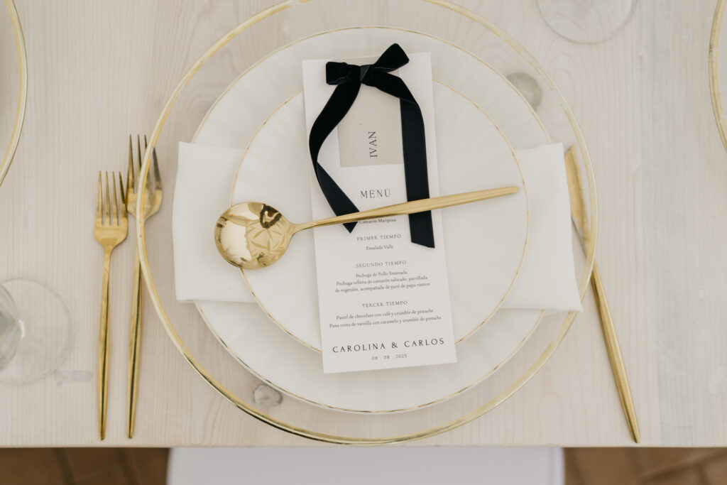 FOTOGRAFIA DEL MONTAJE EN DONDE SE VEN LOS CUBIERTOS, MENU, PLATO Y BAJO PLATO DE LA DECORACION DE BODA EN VENTANA AL MAR VENUE