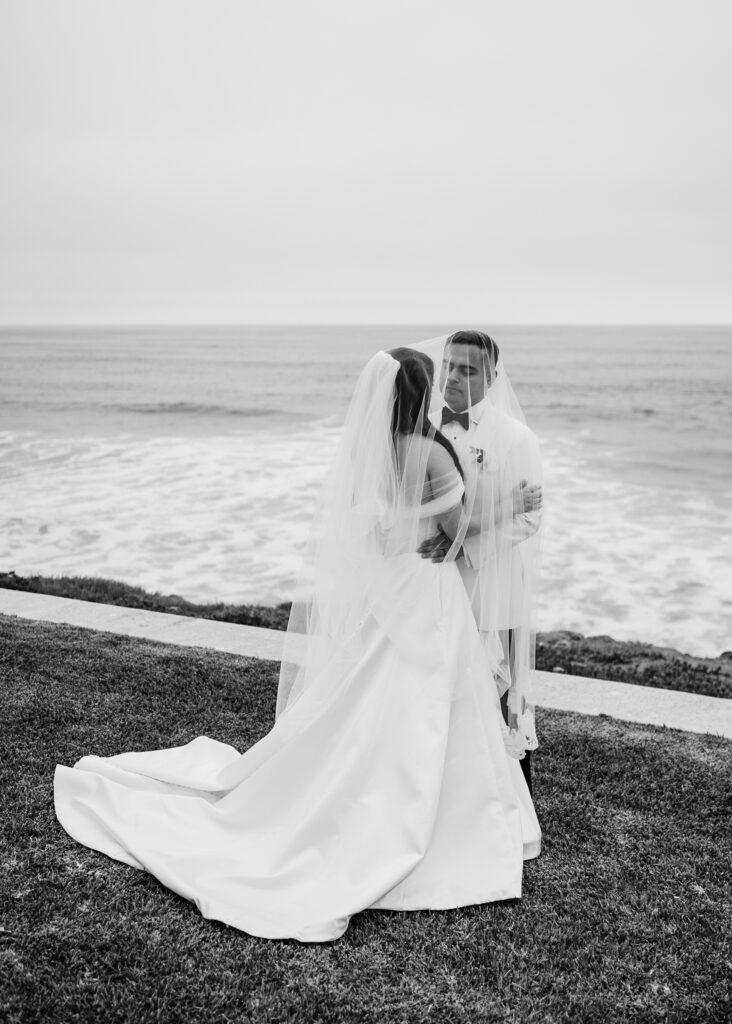FOTOGRAFIA DE NOVIOS CUBIERTOS POR EL VELO FRENTE AL MAR EN VENTANA AL MAR VENUE EN BLANCO Y NEGRO